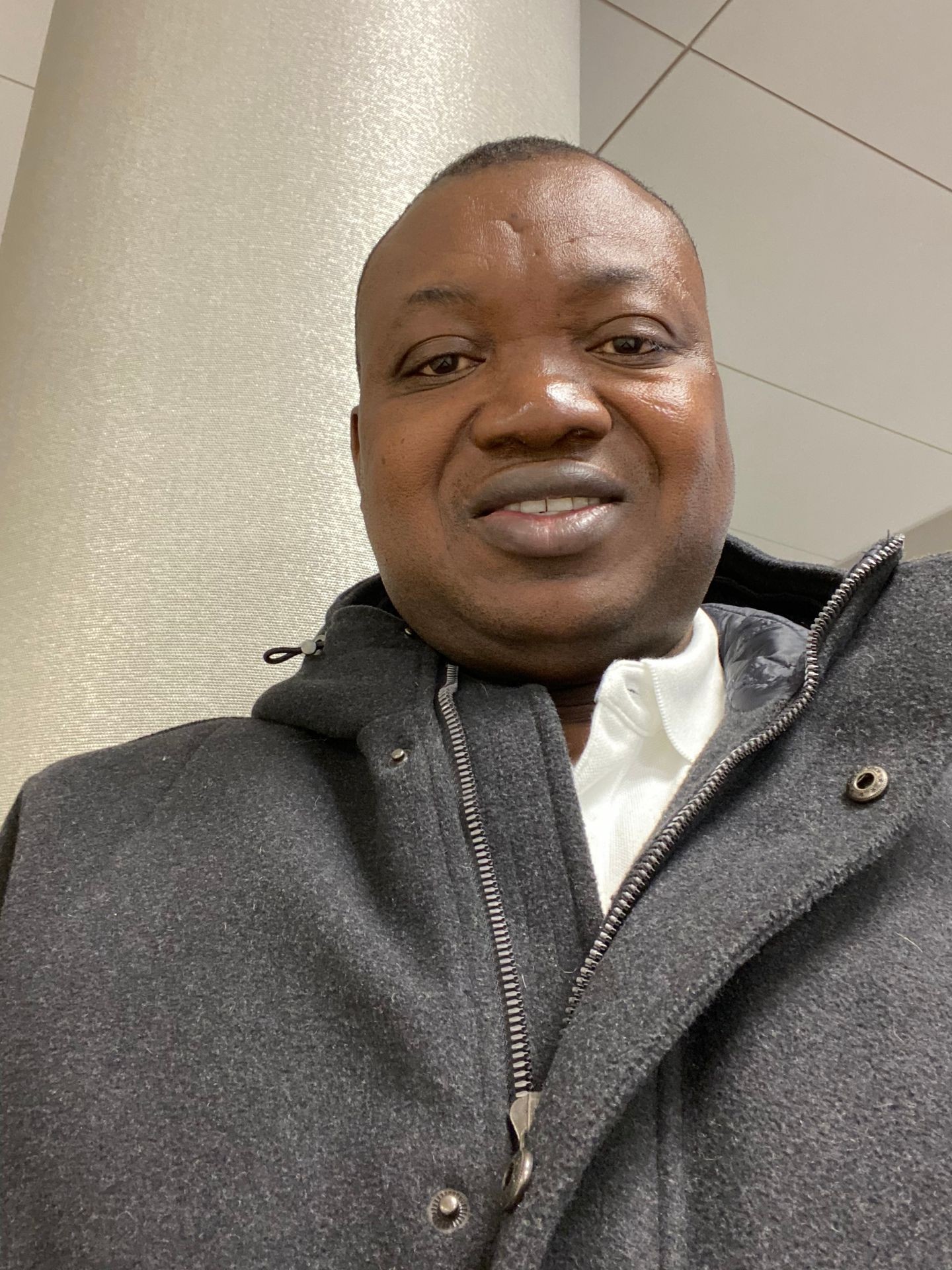 Person in a gray coat smiles, standing in front of a light-colored wall indoors.