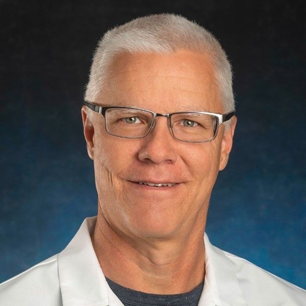 Person with white hair wearing a white lab coat against a dark background.